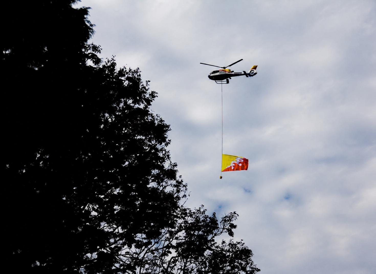 A helicopter carrying the Bhutanese flag above silhouetted trees